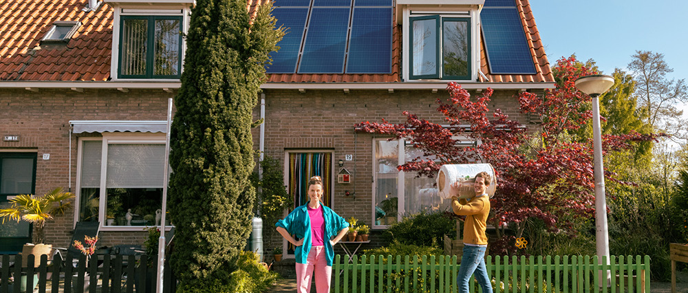 Vrouw staat met handen in de zij voor een huis met zonnepanelen. Er komt een man met een rol isolatiemateriaal aanlopen.