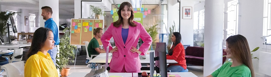 Vrouw staat in roze pak met armen over elkaar aan bureau. Vier andere mensen in gekleurde kleding zitten achter hun bureau achter een computer