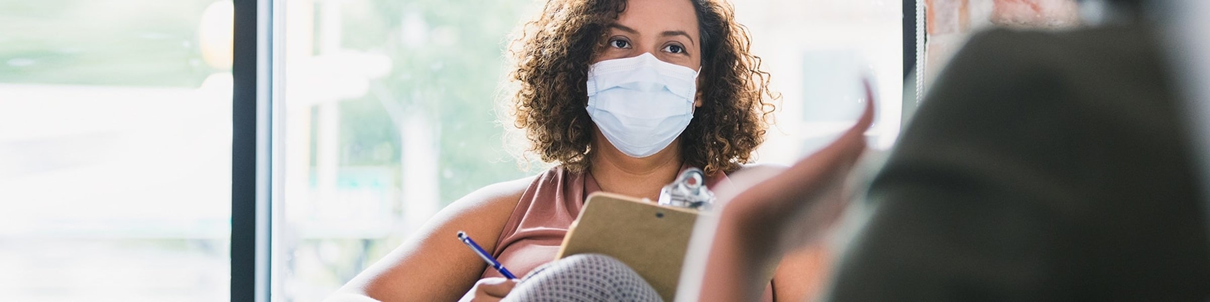 Twee vrouwen zitten tegenover elkaar, een van de vrouwen draagt een mondkapje en maakt aantekeningen