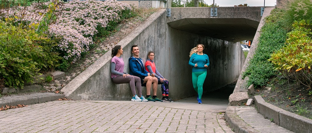 Drie jongeren doen aan wall-sit bij de uitgang van een tunneltje. Een jonge vrouw in sportkleding komt hardlopend het tunneltje uit. | a.s.r. doet het