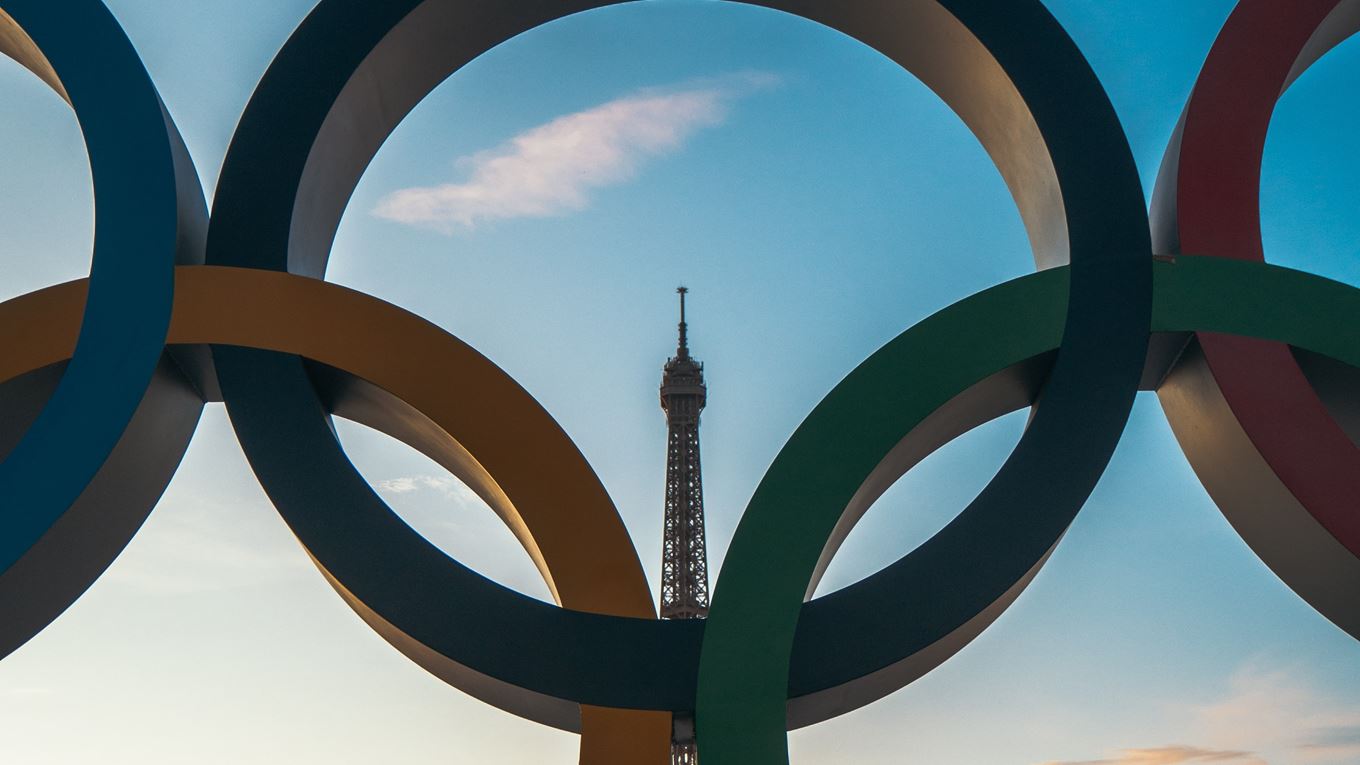 Op de foto zijn de vijf Olympische ringen te zien. Door de zwarte ring heen zie je de punt van de Eiffeltoren