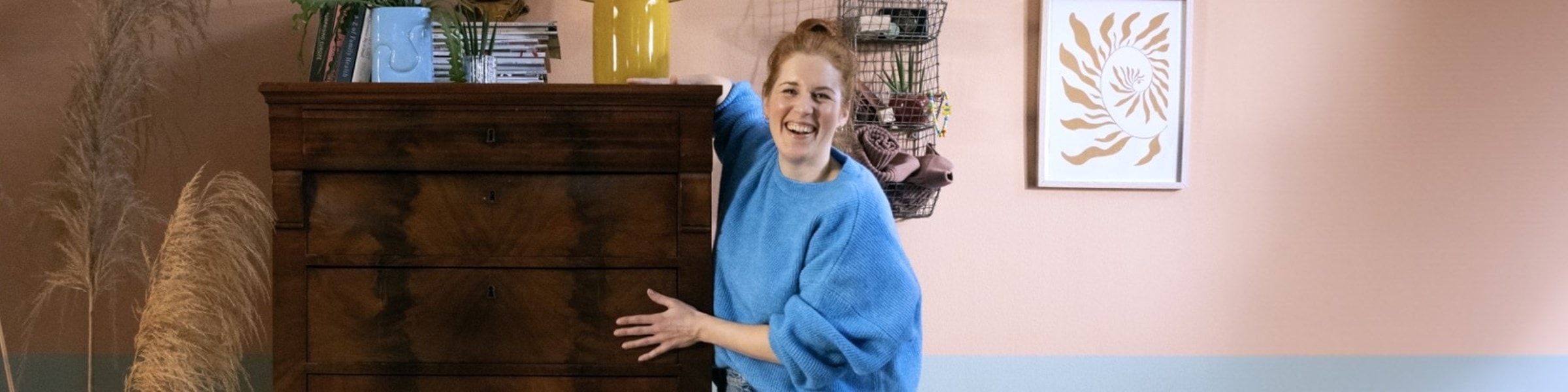 Vrouw staat lachend naast een gerepareerde kast. Op de kast staat een plantje en vaas en op de muur achter haar hangen bloemen en een schilderij.