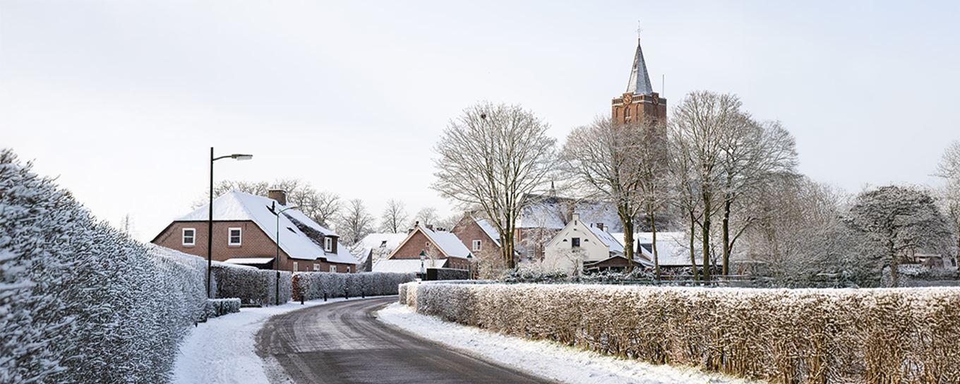 Een wegdek met smeltend sneeuw erop en ook gedeelte niet en met op de achtergrond een dorp met nog witte daken