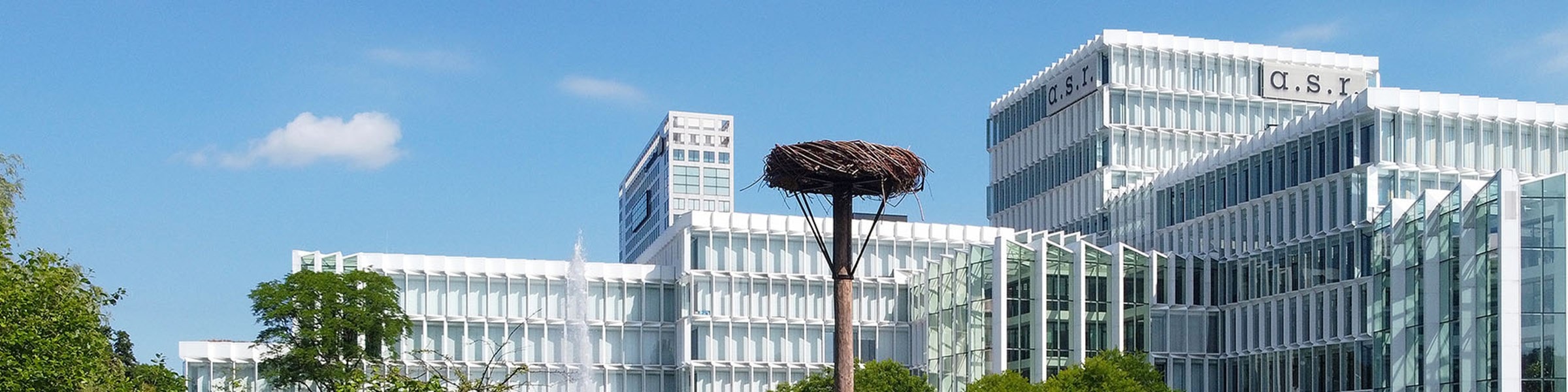 Op de foto staat het gebouw van a.s.r. Hiervoor is een paal met daarop een nest waar ooievaars gebruik van kunnen maken. Het gebouw is wit met veel ramen en op het gebouw staat het logo van asr