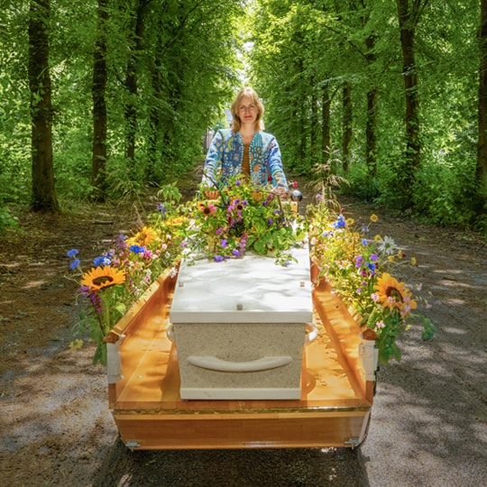 Vrouw fietst door het bos op een uitvaartfiets. De kist is versierd met kleurrijke bloemen.